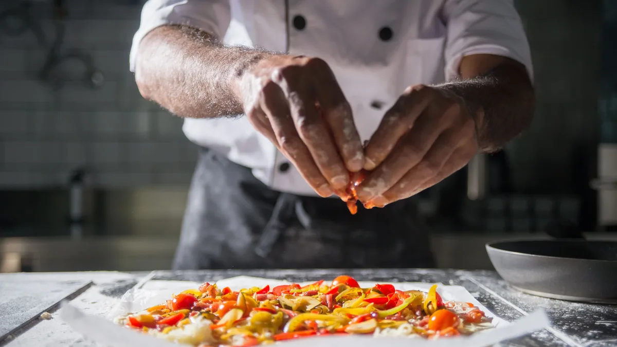 A chef preparing a dish in a kitchen, with the chef's hands visible as they sprinkle something onto a plate of food.