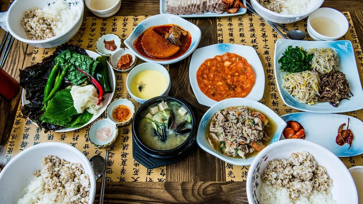 A table laden with an assortment of dishes, including bowls of rice, vegetables, and meat, accompanied by various sauces and condiments, set against a backdrop of a wooden table with a yellow placemat featuring Chinese characters.
